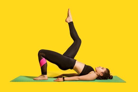 Fitness girl in tight sportswear practicing yoga, doing one legged bridge pose with leg raise, training muscles, flexibility exercises. indoor studio shot, sport workouts isolated on yellow background