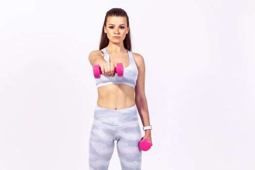 Confident assertive sports woman in white top and tights doing sports exercises holding dumbbells in hands, pumping up muscles, developing strength. Indoor studio shot isolated on gray background