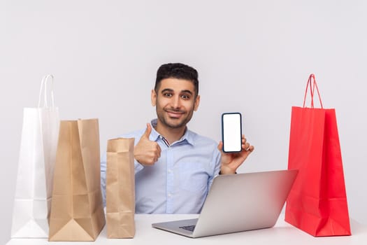 Happy man sitting at desk with laptop and shopping bags around, showing thumbs up, holding cell phone empty screen, mock up display for advertisement. indoor studio shot isolated on white background