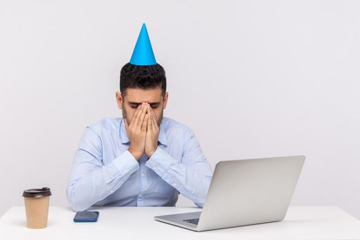 Upset depressed employee guy with party cone on head crying at workplace, hiding face feeling desperate lonely on birthday has to stay in office. indoor studio shot isolated on white background