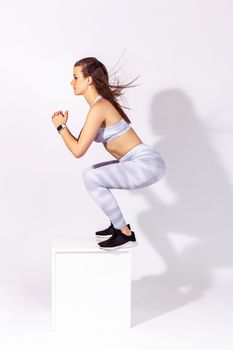 Full length side view assertive fitness woman jumping on cube training muscles and developing strength, working out to be slim and healthy. Indoor studio shot isolated on gray background