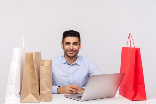 Positive handsome man sitting at desk with laptop, surrounded by packages, looking at camera smiling satisfied with internet shopping, purchase in online store. studio shot isolated, white background