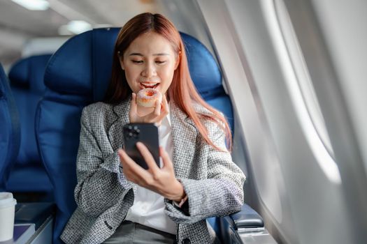 portrait of A successful asian businesswoman or female entrepreneur in formal suit in a plane sits in a business class seat and eating and using smartphone during flight.