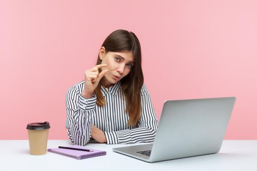 Unhappy frustrated woman office worker sitting at workplace with laptop showing a little bit gesture, measuring minimum talking about her salary. Indoor studio shot isolated on pink background