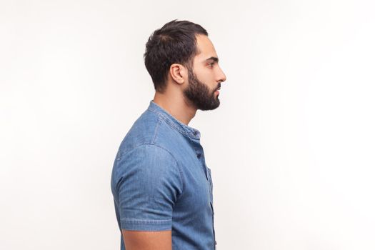 Profile portrait of serious self assured unsmiling man with beard in blue shirt seriously looking far away, confidence. Indoor studio shot isolated on white background