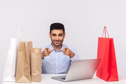 Cheerful handsome man sitting at desk with laptop, surrounded by packages, pointing to camera smiling, recommending internet shopping, purchase in online store. studio shot isolated, white background