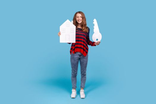Full length portrait of smiling woman wearing sweater, holds big key and paper house, looking at camera with smile, rejoicing purchase of real estate Indoor studio shot isolated on blue background.