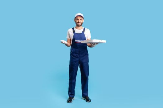 Full length happy courier in workwear and protective gloves carrying pizza box, holding empty space on palm for advertise image. Fast food delivery service. studio shot isolated on blue background
