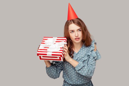 Charming brunette girl with funny party hat holding gift listening to box, curious what bonus inside, celebrating birthday, holiday event, festive mood. studio shot isolated on gray background