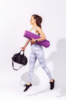 Side view full length energetic athletic woman in white sportswear holding bag and fitness mat hurrying like flying gym to workout and pump up muscles. Indoor studio shot isolated on gray background