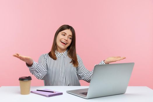 Kind open hearted woman office worker sitting at workplace looking at laptop screen on video call with raised arms, wants to hug or share something. Indoor studio shot isolated on pink background