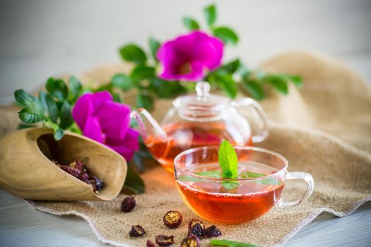 brewed rosehip tea in a glass teapot with rosehip flowers and mint, on a wooden table.