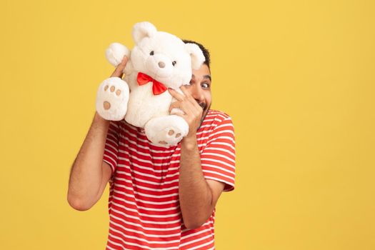 Funny playful man in striped t-shirt having fun hiding behind white fluffy teddy bear, making present, wishing happy holidays. Indoor studio shot isolated on yellow background