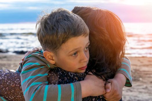 a cute sad preschooler boy hugs his mother on the seashore in the sunset light. love for parents. Close family relationships