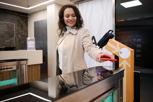 Charming successful Hispanic young woman placing her smartphone over QR code reader at check-in counter while entering to a VIP lounge in the airport departure terminal during her business trip