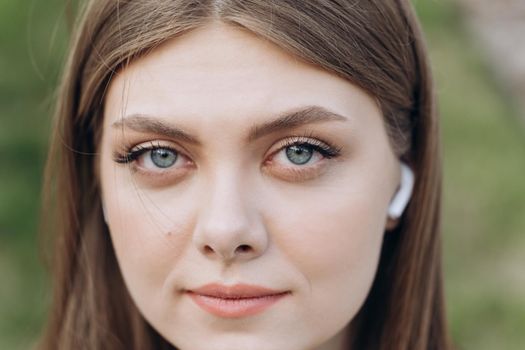 Face of beautiful ukrainian woman with wireless earphones looking at camera. Portrait young caucasian pretty brunette girl outside at sunshine. Etnnicity. People. Beauty.