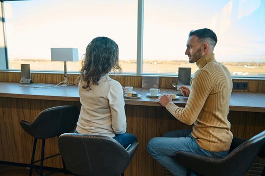 Young married Caucasian couple having a snack together in an airport restaurant, sitting by the panoramic windows overlooking the runway, waiting to board the flight during their honeymoon