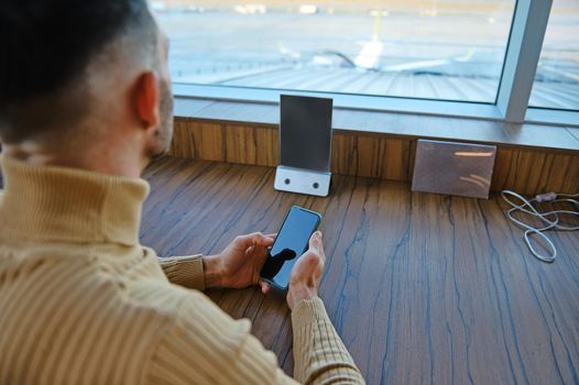 Focus on a smartphone with copy space for advertising text on the blank screen in the hands of a man business traveler, entrepreneur looking at runways in the airport departure terminal