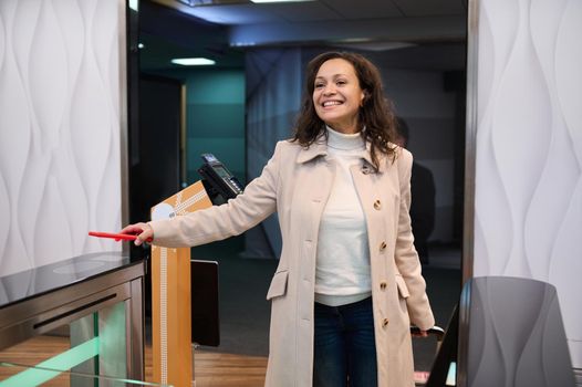 Smiling multiethnic beautiful woman, in casual wear on a business trip, scans information on her smartphone, places it over the QR code reader at the check-in counter in the airport departure terminal