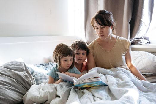 Woman is reading a book for two siblings while they are all sitting in the bed covered with white linen. High quality photo