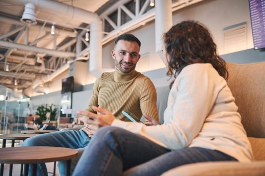 Happy young Caucasian couple talking while sitting in VIP lounge of an airport departure terminal waiting to board flight. Business, travel, journey concept