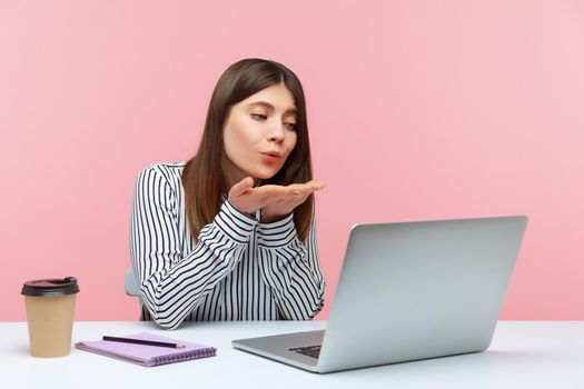 Cute happy brunette woman in striped shirt sending air kisses at laptop display sitting on workplace, flirting chatting via web camera, online date. Indoor studio shot isolated on pink background
