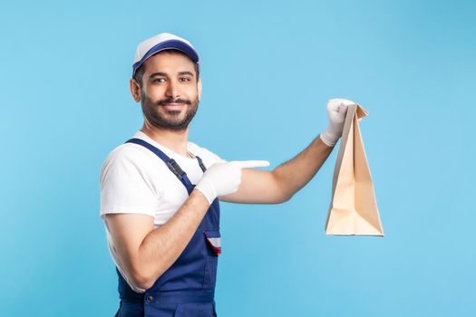 Look at bag. Portrait of cheerful handyman in overalls and gloves pointing at ordered parcel. Courier delivering food in paper package, post mail services. studio shot isolated on blue background