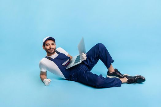 Full length handsome professional handyman in uniform lying sexy, posing on camera and smiling, holding laptop for online customer support, contact center of repair service. studio shot isolated