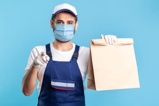 Hey you. Portrait of cheerful bearded handyman in overalls, mask and gloves carrying parcel, pointing to camera. Courier delivering food in paper bag, post mail services. indoor studio shot, isolated