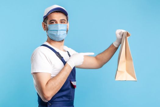 Look at bag. Portrait of cheerful handyman in overalls, mask and gloves pointing at ordered parcel. Courier delivering food in paper package, post mail services. indoor, isolated on blue background