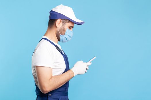 Side view, handyman in overalls and mask typing on mobile phone, using cellphone messenger to accept online order as delivery, repair and maintenance services. indoor shot isolated on blue background