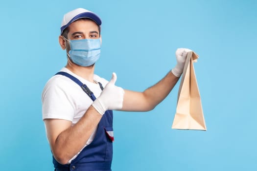 Excellent job. Portrait of satisfied handyman in overalls, mask and gloves holding ordered parcel, showing thumbs up, recommending delivery, post mail service. indoor shot isolated on blue background