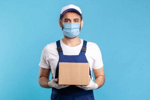 Delivery and cargo transportation. Professional loader in workwear and mask holding cardboard box, smiling to camera. Courier carrying parcel. Online express shipping, relocation moving services.