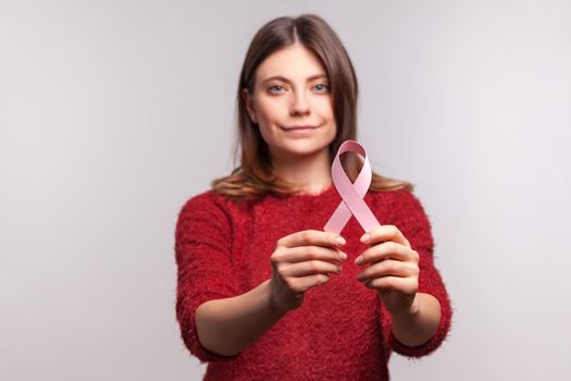 Portrait brunette woman in shaggy sweater showing pink ribbon, symbol of breast cancer awareness, looking at camera with serious responsible expression. indoor studio shot isolated on gray background