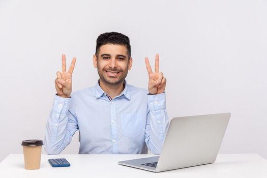 This is win. Joyful businessman sitting office workplace with laptop on desk, showing victory gesture, celebrating successful project, job promotion. indoor studio shot isolated on white background