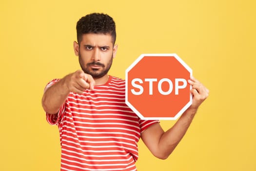 Strict angry man with beard in striped t-shirt holding stop sign and pointing at camera with serious face expression, control, security. Indoor studio shot isolated on yellow background