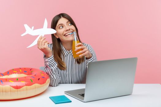 Positive woman drinking lemonade holding paper plane in hand planning vacation abroad sitting at workplace with laptop, passport and rubber ring on desk. Indoor studio shot isolated on pink background