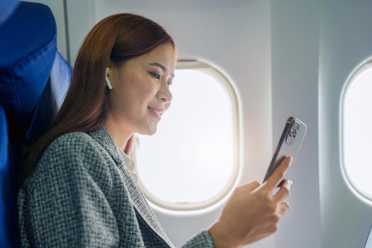 A successful asian businesswoman or female entrepreneur in formal suit in a plane sits in a business class's seat and uses a smartphone during flight.