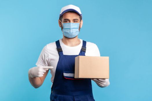 Cargo transportation, delivery service. Friendly worker in uniform and mask pointing at cardboard box and smiling to camera. Courier carrying parcel. Online express shipping, relocation service.