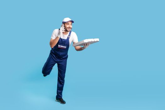 Express delivery service. Full length funny happy courier man in uniform talking on phone and running to give pizza on time, hurrying to deliver fast food. studio shot isolated on blue background