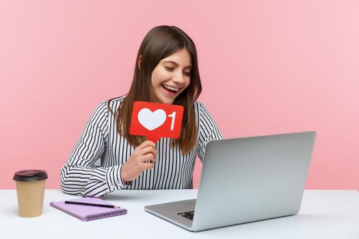 Smiling optimistic woman blogger chatting with followers on video call at laptop asking to rate her posts showing likes counter template. Indoor studio shot isolated on pink background