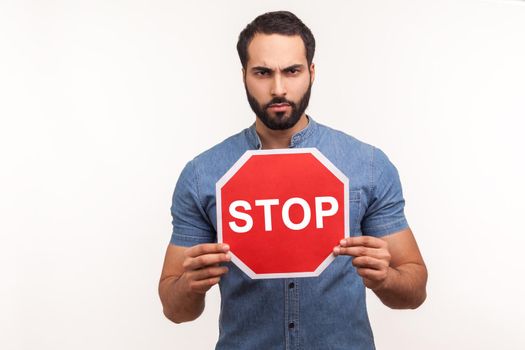Concerned nervous bearded man in blue shirt holding red stop sign looking at camera with serious expression. Indoor studio shot isolated on white background