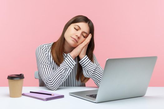 Bored sleepy woman office worker in striped shirt napping leaning head on hands sitting at workplace with laptop, overworking, early awakening. Indoor studio shot isolated on pink background