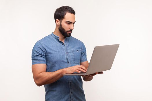Self confident serious man with beard in blue shirt holding wireless computer in hand, working on laptop, making edits, deadline. Indoor studio shot isolated on white background
