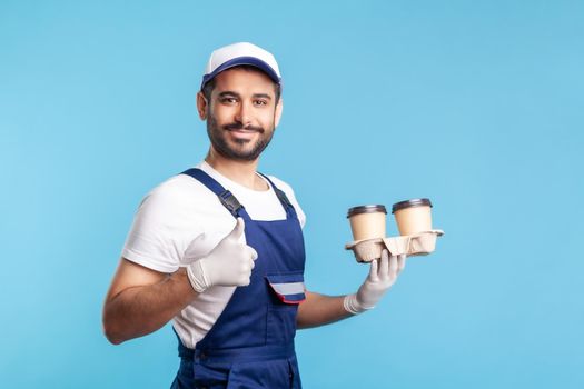 Delivery service. Side view, friendly courier in overalls holding coffee, wearing safety gloves offering drinks in disposable cups and showing thumbs up, like gesture. indoor studio shot, isolated