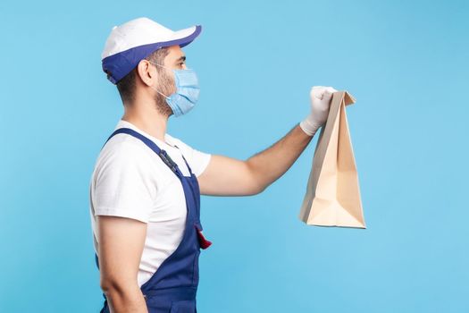 Side view, friendly professional courier in uniform, mask and safety gloves giving groceries bag, delivering takeaway food, post services express shipping. indoor shot isolated on blue background