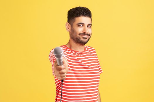 Bearded male reporter in striped t-shirt holding microphone, making interview and asking opinion, discussing important topics. Indoor studio shot isolated on yellow background