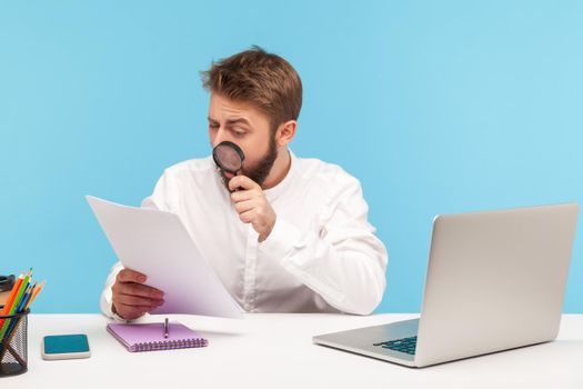 Serious businessman attentively studying analyzing papers holding magnifying glass, looking for mistakes and errors, checking datas sitting at workplace. Indoor studio shot isolated on blue background