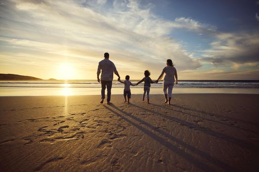 Rear view shot of a carefree family holding hands while walking on the beach at sunset against cloudy sky. Loving parents and their two kids spending time together by the sea watching sunset.