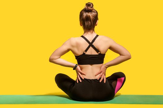 Back view, sportswoman sitting on mat and touching sore spine, suffering backache after physical training or yoga practice, massaging stiff muscles. indoor studio shot isolated on yellow background